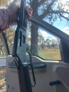 Person holding rifle in open car door outdoors.